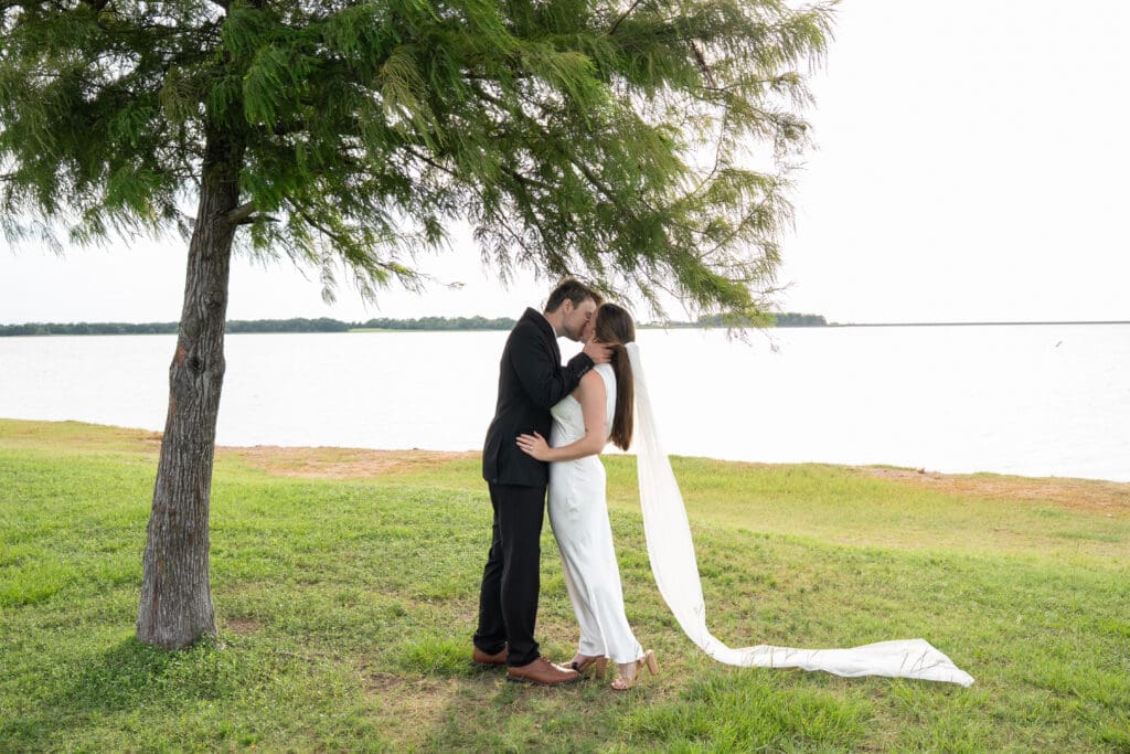 Bride and Groom share their first kiss under a tree at sunset with a lake in the background - when to elope in Texas in Summer